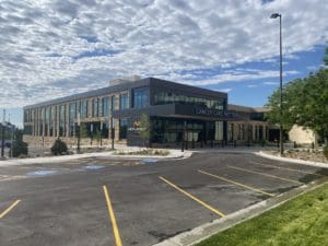 Exterior of the cancer care institute with black paneling and large black windows in the front. Surrounded by trees and a black parking lot with yellow lines.