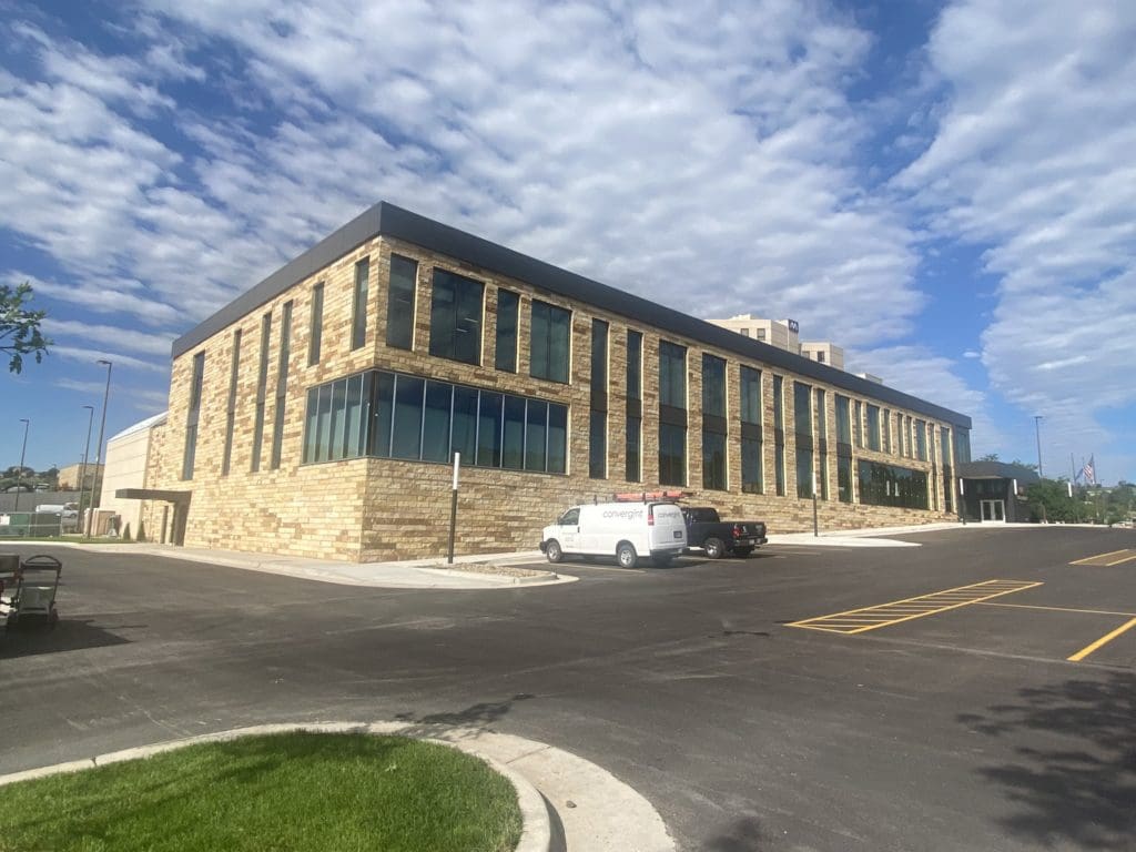 exterior of a white brick building with gray ceiling and large windows on the front and side of the building. Bright blue sky with large white clouds in the background.