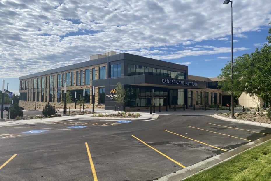 Exterior of the cancer care institute with black paneling and large black windows in the front. Surrounded by trees and a black parking lot with yellow lines.